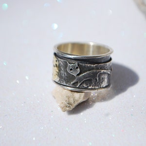 May include: A silver and gold ring with a textured surface and a cat design. The ring is on a white background.