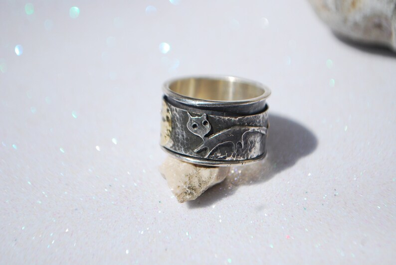 May include: A silver and gold ring with a textured surface and a cat design. The ring is on a white background.