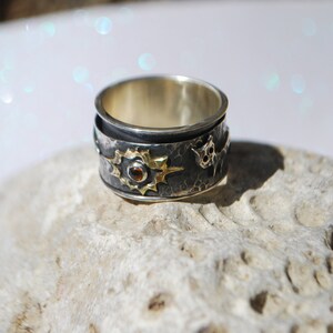May include: A silver and gold ring with a textured band and a star design. The star has a small red gemstone in the centre.