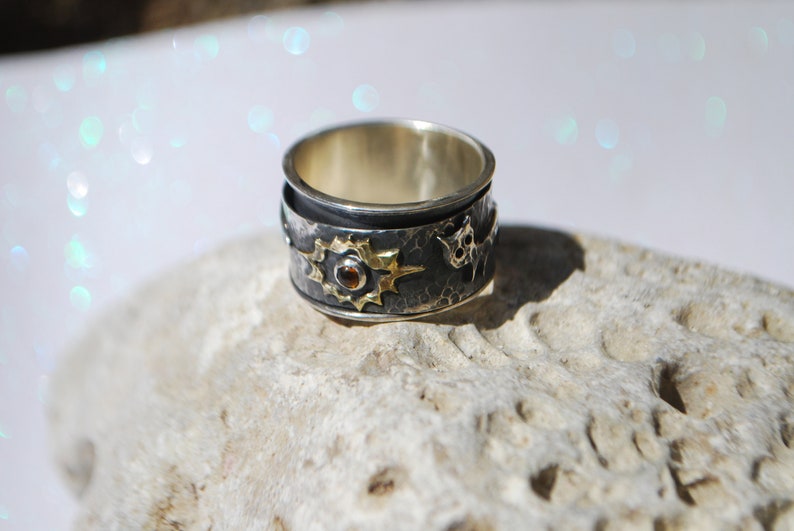 May include: A silver and gold ring with a textured band and a star design. The star has a small red gemstone in the centre.