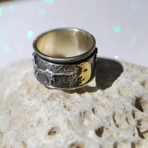 May include: A silver ring with a textured surface and a gold crescent moon design. The ring is set on a white, textured surface.