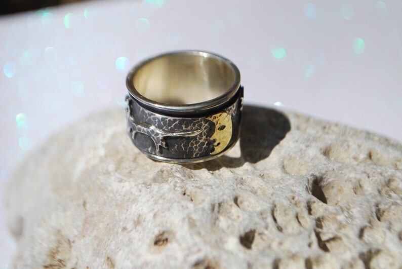 May include: A silver ring with a textured surface and a gold crescent moon design. The ring is set on a white, textured surface.