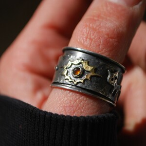 May include: A silver ring with a gold accent and a small orange gemstone. The ring is textured and has a unique design.