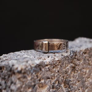 May include: A silver ring with a textured finish and a rectangular silver accent. The ring is sitting on a rough, gray surface.