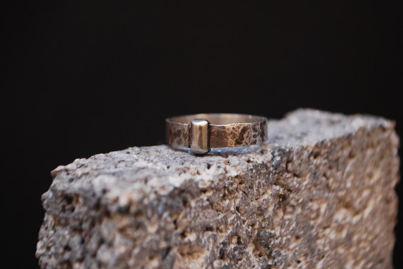 May include: A silver ring with a textured finish and a rectangular silver accent. The ring is sitting on a rough, gray surface.