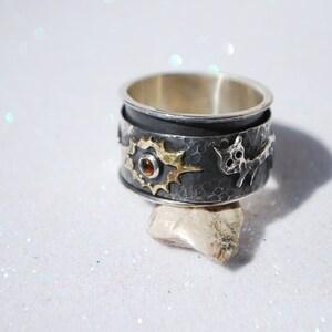 May include: A silver ring with a black oxidised finish, featuring a sun design with a small orange gemstone in the centre, and a stylised animal figure on the side.