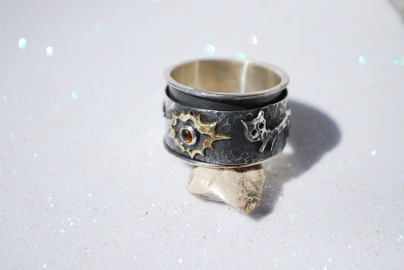 May include: A silver ring with a black oxidised finish, featuring a sun design with a small orange gemstone in the centre, and a stylised animal figure on the side.