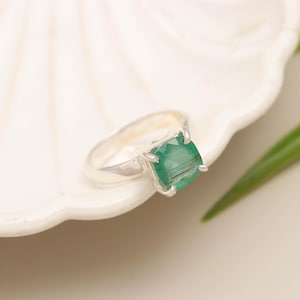 Op de afbeelding: Een zilveren ring met een vierkant geslepen, doorschijnende groene edelsteen in een zetting met vier pootjes. De ringband is glad en gepolijst en toont een eenvoudig metalen ontwerp dat de natuurlijke insluitingen in de steen benadrukt.
