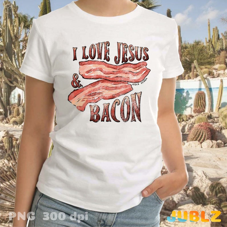 Puede incluir: Camiseta blanca con el texto "I love Jesus & Bacon" en letras rojas y marrones. El texto est&aacute; rodeado por un gr&aacute;fico de dos rebanadas de tocino.