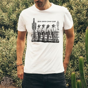 May include: A white t-shirt with a black and white illustration of four cowboys standing in front of a fence with the text "Make America Cowboy Again".
