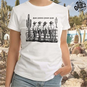 May include: A white t-shirt with a black and white illustration of four cowboys standing in front of a fence with the text "Make America Cowboy Again".
