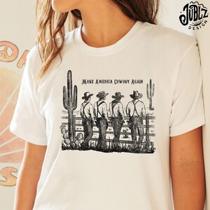 May include: White t-shirt with a black and white illustration of four cowboys standing in front of a fence with cacti. The text "Make America Cowboy Again" is printed on the shirt.