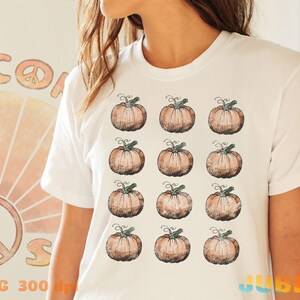 Puede incluir: Camiseta blanca con un patr&oacute;n de 12 calabazas naranjas con tallos verdes. Las calabazas est&aacute;n dispuestas en cuatro filas de tres.