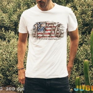 May include: White t-shirt with a distressed American flag design and the text "1776" printed on the front.