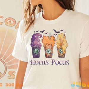 May include: White t-shirt featuring a vintage-style graphic of three Halloween-themed coffee cups with the text "Hocus Pocus" underneath.