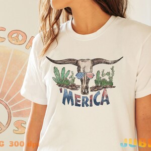 May include: White t-shirt with a distressed graphic of a cow skull wearing sunglasses and surrounded by cacti. The text 'MERICA' is printed below the skull.