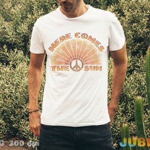 May include: A white t-shirt with a retro-style graphic design featuring a peace sign and the text "Here Comes The Sun".