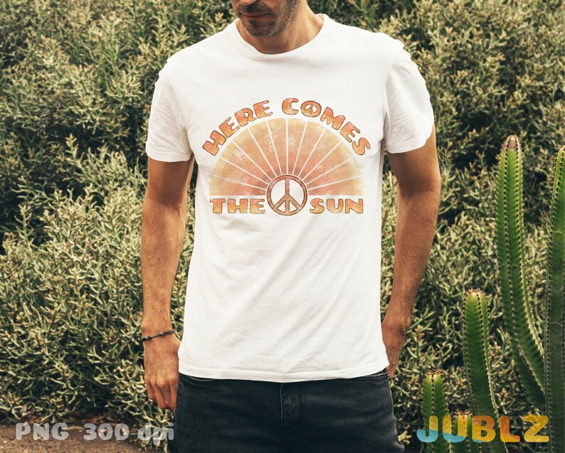May include: A white t-shirt with a retro-style graphic design featuring a peace sign and the text "Here Comes The Sun".