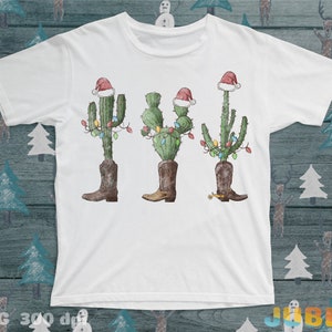 K&ouml;nnte beinhalten: Wei&szlig;es T-Shirt mit einem Weihnachtskaktus-Design. Der Kaktus tr&auml;gt einen Weihnachtsmann-Hut und hat Weihnachtslichter um sich herum. Der Kaktus tr&auml;gt Cowboystiefel.