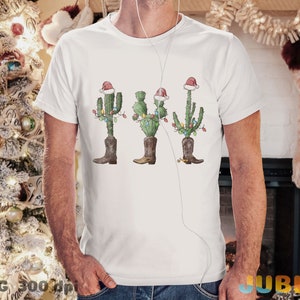 K&ouml;nnte beinhalten: Wei&szlig;es T-Shirt mit einem Weihnachtskaktus-Motiv. Der Kaktus tr&auml;gt einen Weihnachtsmann-Hut und Stiefel und ist mit Weihnachtslichtern geschm&uuml;ckt.