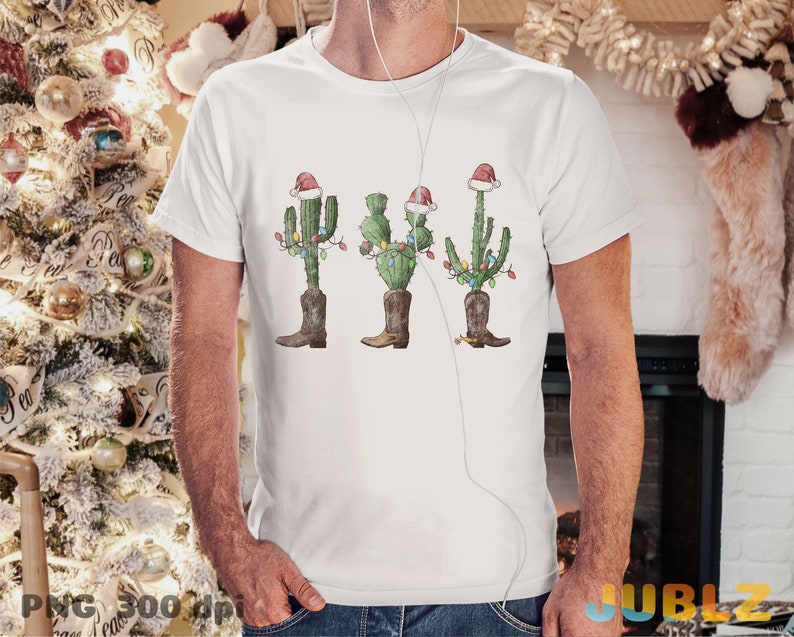 K&ouml;nnte beinhalten: Wei&szlig;es T-Shirt mit einem Weihnachtskaktus-Motiv. Der Kaktus tr&auml;gt einen Weihnachtsmann-Hut und Stiefel und ist mit Weihnachtslichtern geschm&uuml;ckt.