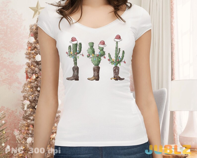 K&ouml;nnte beinhalten: Wei&szlig;es T-Shirt mit einem Weihnachtskaktus, der einen Weihnachtsmann-Hut und Stiefel tr&auml;gt. Der Kaktus ist mit bunten Lichtern geschm&uuml;ckt.