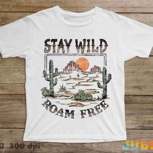 May include: White t-shirt with a vintage-style graphic design. The design features a desert landscape with cacti, a setting sun, and the text "Stay Wild Roam Free".