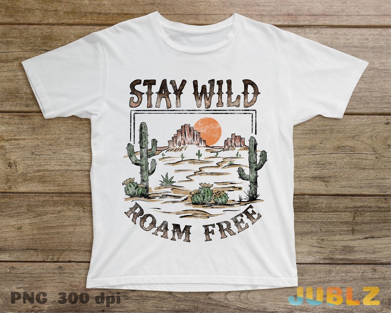 May include: White t-shirt with a vintage-style graphic design. The design features a desert landscape with cacti, a setting sun, and the text "Stay Wild Roam Free".