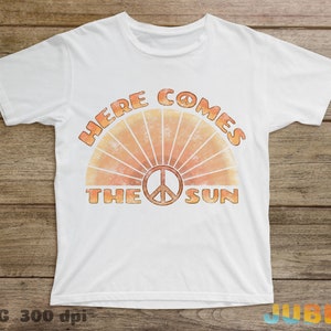 May include: White t-shirt with a retro-style graphic design featuring a peace sign and the text "Here Comes The Sun".