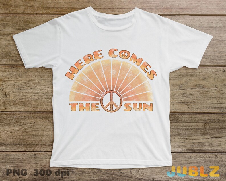 May include: White t-shirt with a retro-style graphic design featuring a peace sign and the text "Here Comes The Sun".