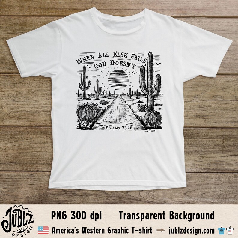 May include: A white t-shirt with a black and white graphic design of a desert landscape with cacti and a road. The text "WHEN ALL ELSE FAILS GOD DOESN'T" is written in a circle around a sun. The text "PSALMS 73:26" is written below the graphic.