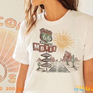 May include: White t-shirt with a vintage Route 66 Motel graphic. The graphic features a retro sign with the text "Route 66 Vacancy Motel" and a sun with rays. The graphic also includes a desert landscape with cacti and a road.