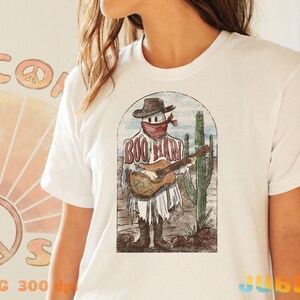 May include: White t-shirt with a vintage-style illustration of a ghost wearing a cowboy hat and bandana, playing a guitar in a desert landscape. The text "BOO MAN" is written on the ghost's shirt.