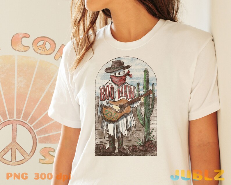 May include: White t-shirt with a vintage-style illustration of a ghost wearing a cowboy hat and bandana, playing a guitar in a desert landscape. The text "BOO MAN" is written on the ghost's shirt.