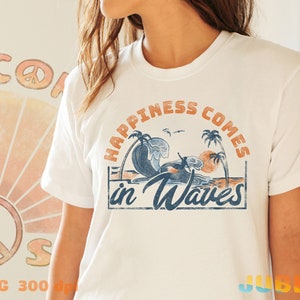 May include: White t-shirt with a vintage-style graphic that says "Happiness comes in waves". The graphic features a sunset, palm trees, and a surfer riding a wave.