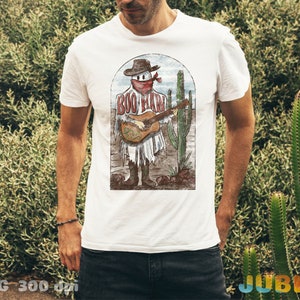 May include: White t-shirt with a vintage illustration of a skeleton playing a guitar in a desert landscape. The skeleton is wearing a cowboy hat and bandana. The text "Boo Haw" is printed on the shirt.