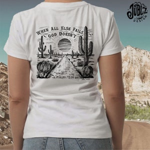 May include: White t-shirt with a black and white illustration of a desert landscape with cacti and a road. The text "When all else fails God doesn't" is above the illustration. The text "Psalms 73:26" is below the illustration.