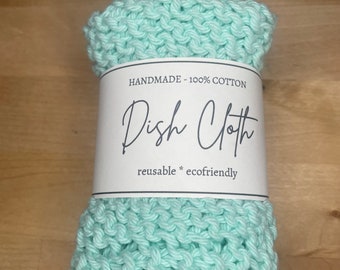 May include: A light green knitted dishcloth made from 100% cotton. The dishcloth is rolled up and has a white label that says "Handmade - 100% Cotton Dish Cloth reusable * ecofriendly".