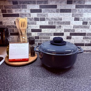May include: A dark blue cast iron Dutch oven with a matching crocheted lid cover sits on a countertop. A wooden spoon and spatula are in a glass jar. A recipe card and a pepper grinder are also visible.