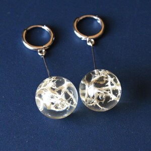 May include: A pair of silver hoop earrings with clear, spherical pendants. Each pendant contains a white, web-like design. The earrings are displayed against a dark blue background, showcasing the jewelry's delicate details.