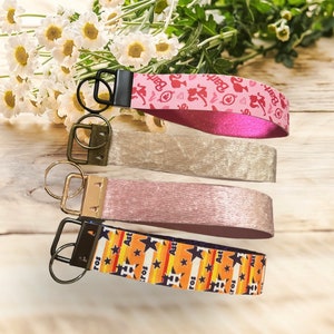 May include: Four keychains with different designs. The top keychain is pink with a white background and the text "Barbie" repeated. The second keychain is a light beige color. The third keychain is a light pink color. The bottom keychain is orange and black with stars and the text "Art" repeated.