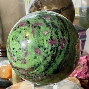 Könnte beinhalten: Eine polierte Kugel aus grünem Zoisit mit Rubin-Einschlüssen, die auf einem transparenten Acrylständer ruht. Der Edelstein weist ein wirbelndes Muster aus grünen, schwarzen und roten Farbtönen auf. Weitere dekorative Kugeln und Kristalle sind im Hintergrund zu sehen.