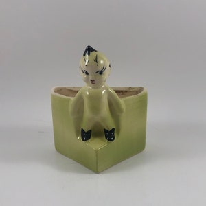 Könnte beinhalten: Ein grüner Keramik-Pflanztopf in Dreiecksform mit einer kleinen Figur eines sitzenden Menschen auf der Oberseite. Die Figur trägt ein gelbes Outfit mit schwarzen Schuhen und hat schwarze Haare und ein weißes Gesicht.