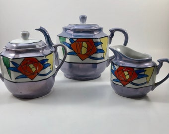 Puede incluir: Un juego de té vintage con un diseño floral geométrico morado, blanco, amarillo, azul y rojo. El juego incluye una tetera, una jarra de crema y un azucarero.