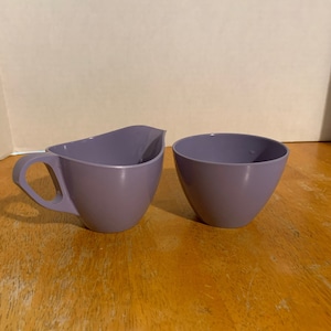 May include: A set of two purple ceramic creamer and sugar bowls. The creamer has a handle and a rounded shape. The sugar bowl is round and has a smooth surface.