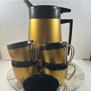 May include: A set of gold and black coffee mugs with a matching gold and black coffee pot. The mugs have gold handles and a black band around the top. The coffee pot has a black lid and handle.