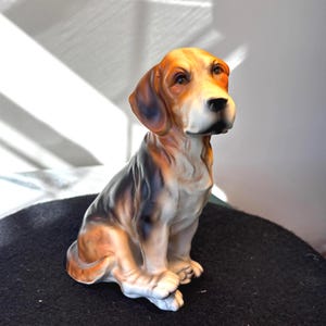 May include: A ceramic figurine of a beagle dog in a sitting position. The dog features a tri-colour coat with patches of brown, black, and white. It has long, drooping ears, a dark nose, and a smooth, glossy finish on its detailed surface.