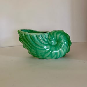 May include: A green ceramic seashell planter. The planter is shaped like a nautilus shell with a smooth, glossy finish.