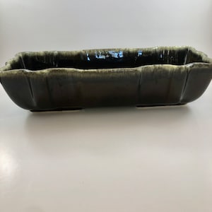 May include: A long, rectangular ceramic planter with a dark green glaze and a light green glaze on the edges. The planter has a slightly wavy rim and four small feet.