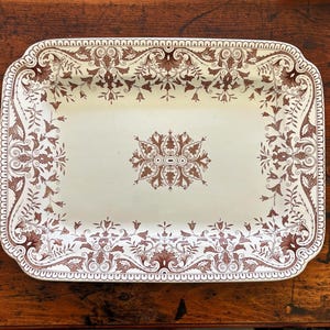 May include: Rectangular serving platter with a cream-colored background and a brown floral pattern. The platter has a decorative border and a central floral design. The edges are slightly scalloped.